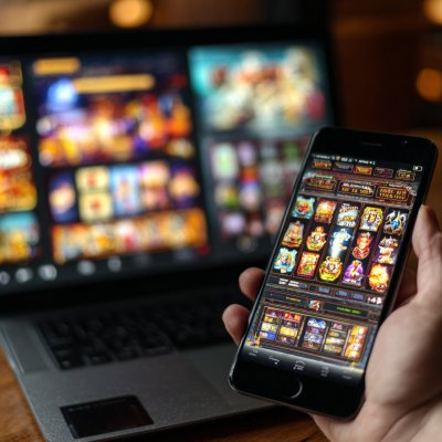 A person holding a smartphone showing a casino slot game, with a laptop in the background also showing the game.